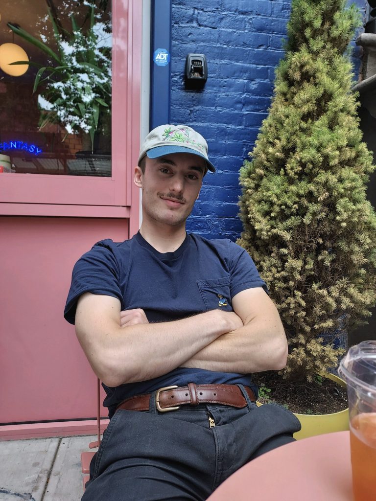 A portrait of a young man in jeans, a navy blue t shirt, and a light blue baseball cap sitting with his arms crossed at an outdoor table