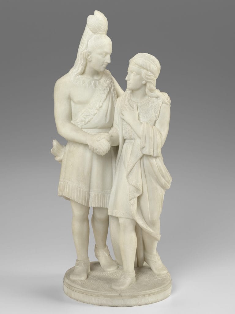 A photo of a white marble sculpture of two Native American figures—a young adult man with long hair and a feathered headpiece and a young woman with long hair and a headband—standing close together and holding hands, gazing at each other with gentle expressions.