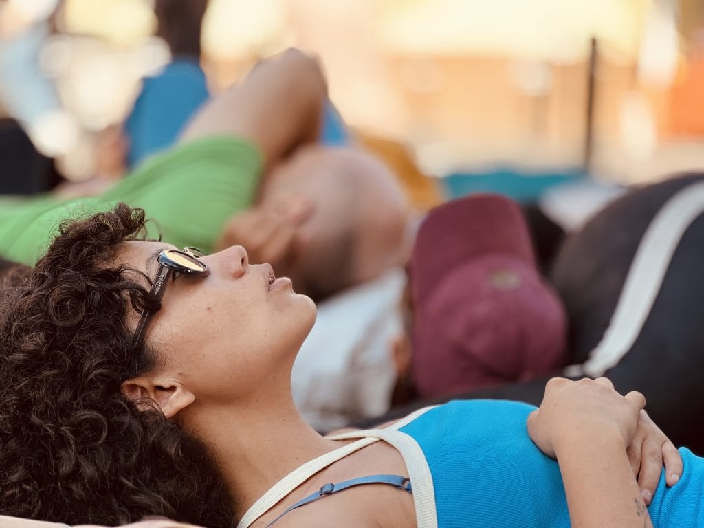 A performer with short curly hair and sunglasses lies on their back outdoors at sunset, wearing a blue tank top, with other performers blurred in the background.