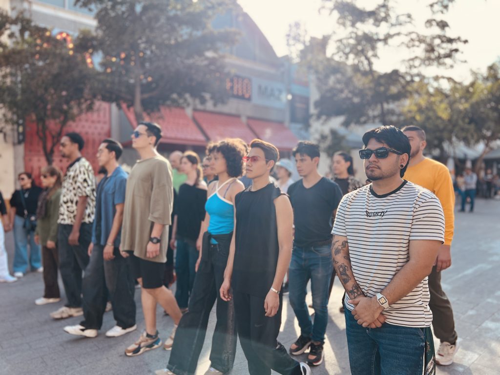 A group of performers stand and walk in formation outside a contemporary museum building in daylight, dressed casually and facing forward.