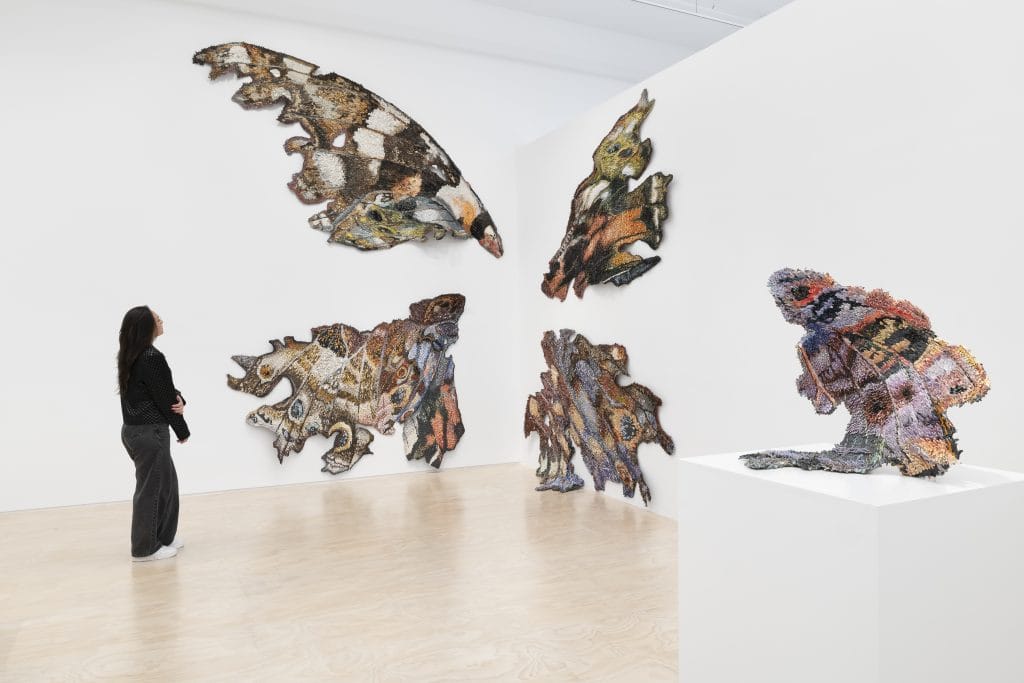 A visitor stands in a bright white gallery viewing a large, irregularly shaped, multicolored sculpture arranged on wall and pedestal, with an additional densely textured sculptural piece displayed on a white pedestal in the foreground. Both artworks by artist Rebecca Manson.