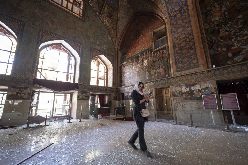 Woman walks through grand historic hall with murals, broken glass scattered across floor, documenting damage.
