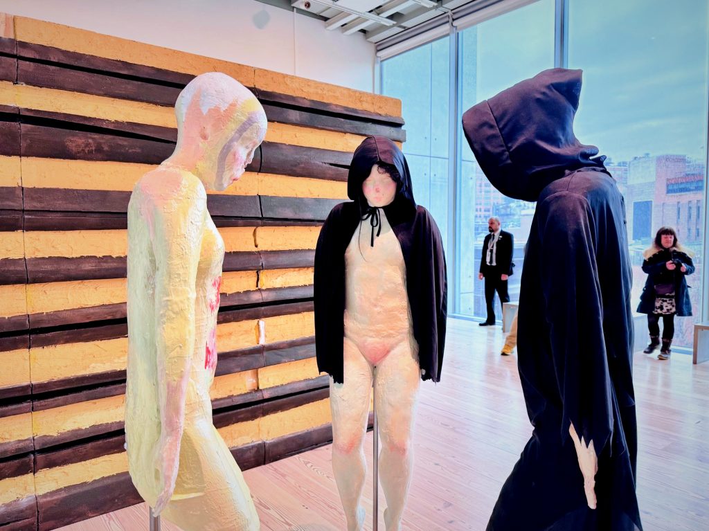 An installation view of Isabelle Frances McGuire’s sculptures at the Whitney Biennial, featuring three life-sized, textured figures, two wearing black hooded cloaks, in a gallery.