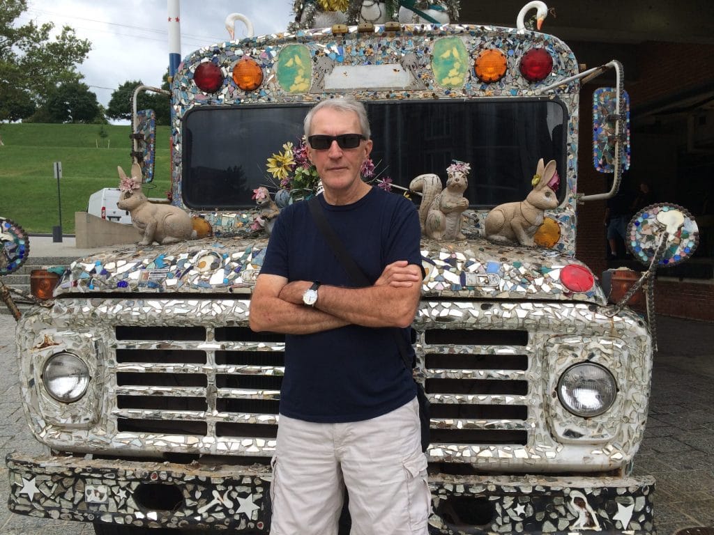 A photo of an older white man, likely in his 60s, wearing dark sunglasses, a navy T-shirt, and light-colored shorts, standing with his arms crossed in front of a mosaic-covered truck decorated with mirrors, colorful tiles, flowers, and small animal sculptures, including rabbits and squirrels.