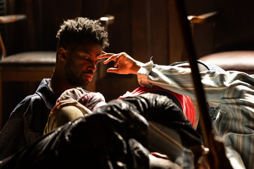 A man with dark, curly hair sits in low light while another person’s hand gently reaches out to touch his forehead amidst a huddle of figures.