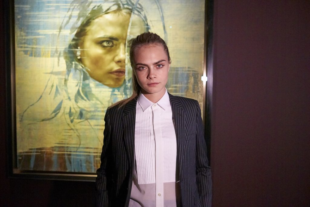 Cara Delevingne stands before her painted portrait by Jonathan Yeo inside gallery exhibition setting.