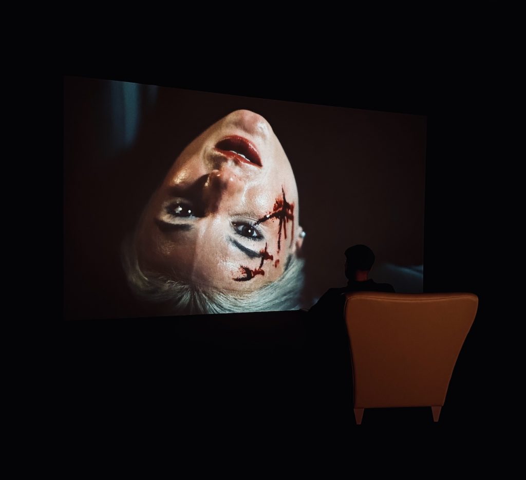 A person in a high-back chair sits in a dark room, silhouetted against a large screen displaying an upside-down, blood-splattered face of a blonde woman.