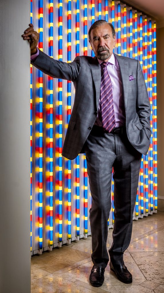 Jorge Pérez poses in a gray suit with pink tie in front of colorful vertical light installation, showcasing his bold personal style and dedication to contemporary art collecting.