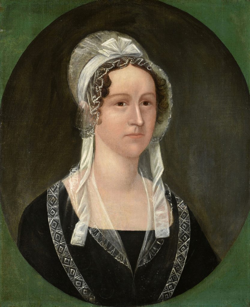 An early 19th-century oval portrait of a woman shown from the chest up against a dark background with a green border. She wears a white bonnet with lace trim and long ribbons framing her face, and a black dress with patterned straps and a sheer white neckline. Her expression is calm and reserved as she faces slightly to the viewer’s left in a formal folk-art style.