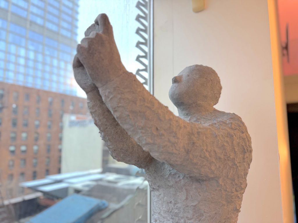 Textured, pale sculpture by Judith Hopf of a human figure holding a smartphone upward, positioned near a window overlooking a blurred urban street scene.