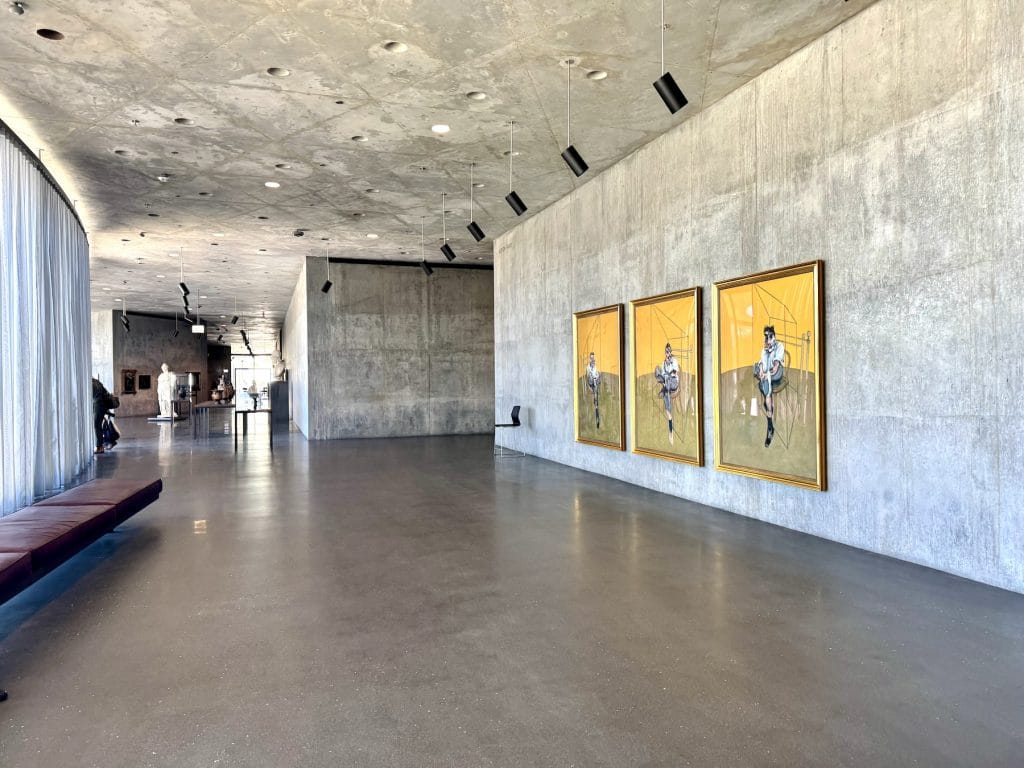 An orange and brown triptych by Francis Bacon is installed in a concrete-walled gallery.