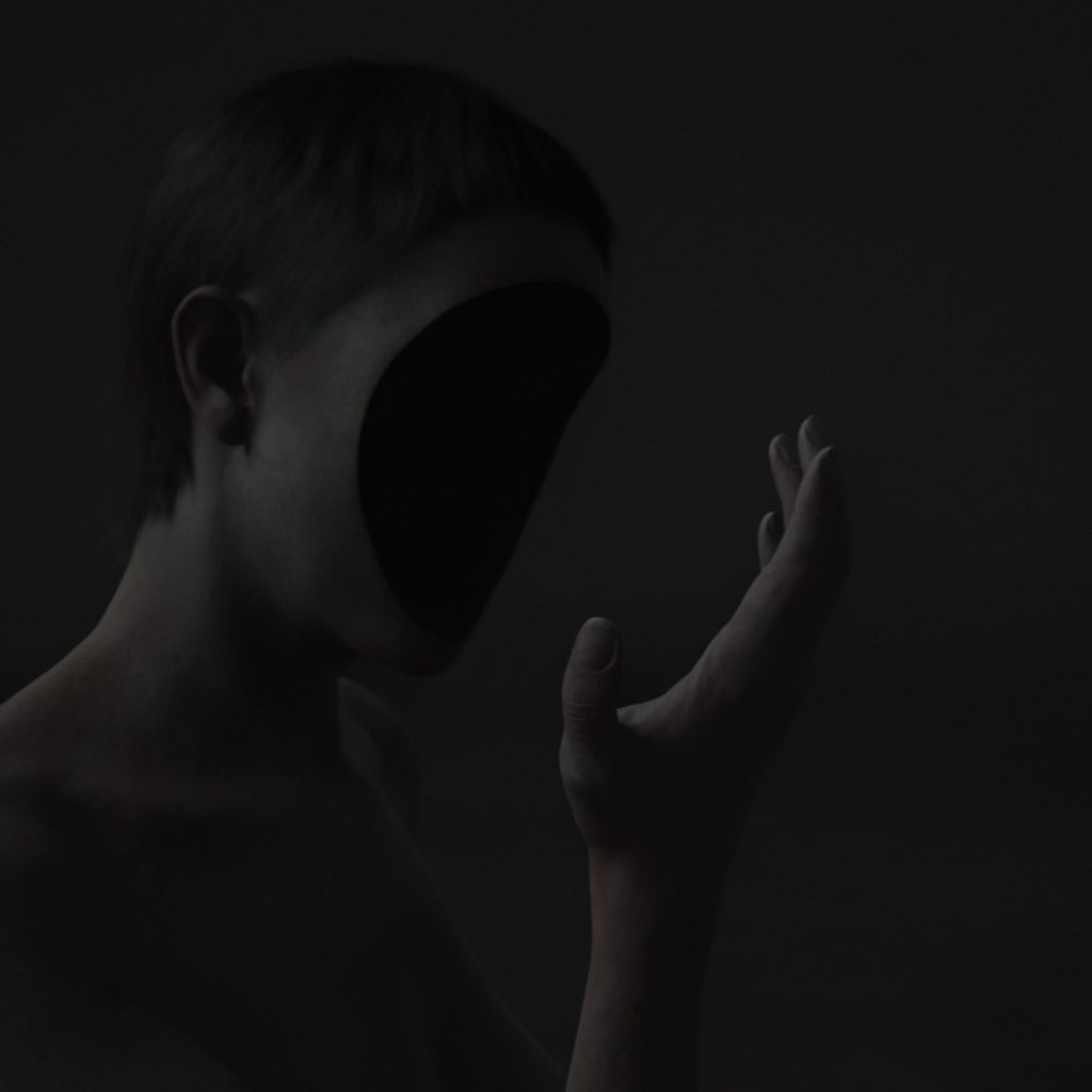 A dimly lit figure with a smooth, featureless black void where the face should be raises a hand against a dark background.