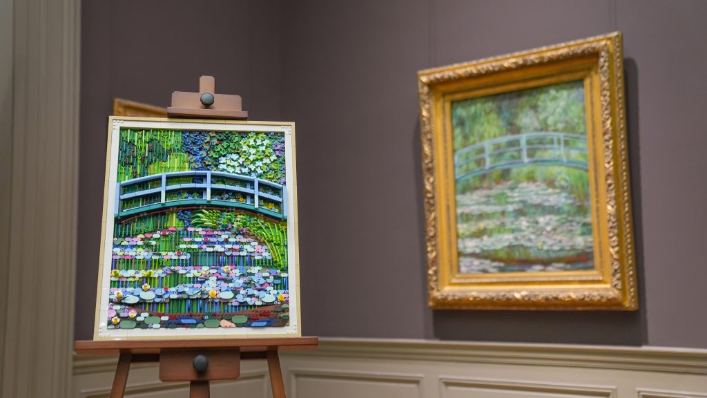 A photo of a colorful LEGO mosaic artwork depicting a curved green bridge over a pond filled with stylized lily pads, displayed on an easel inside a museum gallery, with a framed impressionist painting of the same scene hanging on the wall behind it.