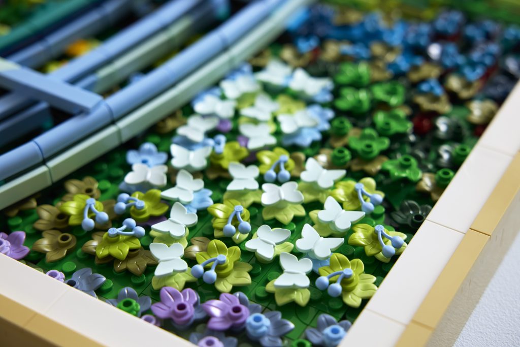 A photo of a close-up detail of a LEGO mosaic artwork showing clusters of butterfly-shaped LEGO pieces in pale blue and white among layered green leaves, with small rounded blue elements resembling cherries scattered throughout, evoking dense foliage beneath a pale green arched bridge and emphasizing the intricate, textured construction.
