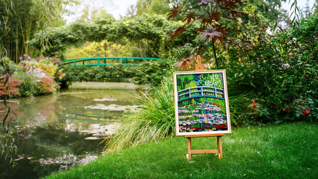 A photo of a LEGO mosaic artwork on a small wooden easel set outdoors beside a calm garden pond, showing a green arched bridge over water dotted with lily pads, with lush plants, grasses, and trees filling the background.