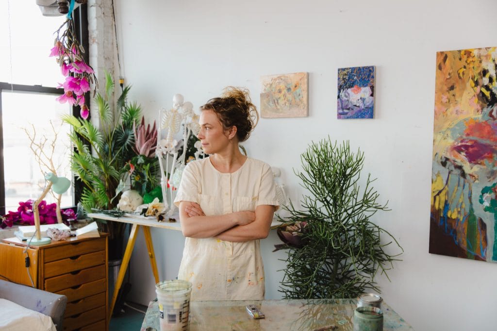 Color photo of a woman artist standing with arms crossed in a light-filled studio with plants, paintings, and art supplies.