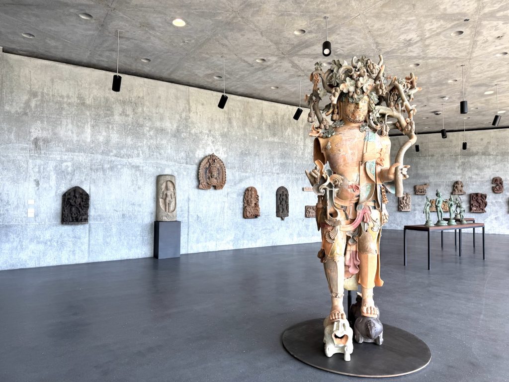 A tall, terracotta-colored patchwork sculpture of a figure with an ornate, branching headdress stands in a concrete gallery surrounded by smaller ancient stone relief carvings.