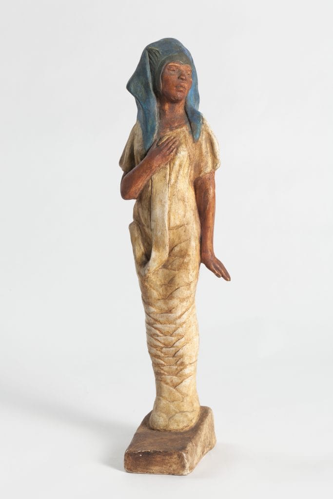 A photo of a small painted sculpture of a young Black woman with a serene expression, wearing a blue Egyptian pharoah-like head covering and a simple cream-colored dress, her right hand resting over her chest as her lower body tapers into a wrapped, column-like form.