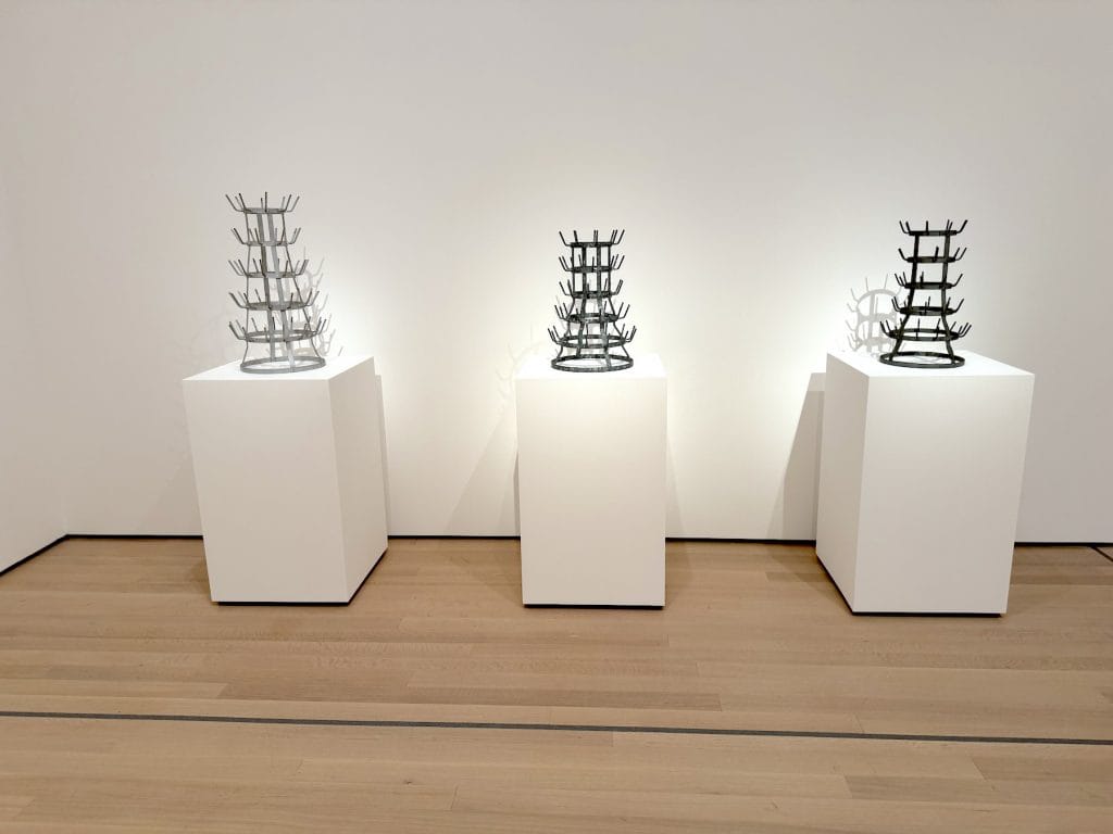 Three different metal bottle rack sculptures by Marcel Duchamp, each displayed on an individual white pedestal in a minimalist gallery with wood floors.