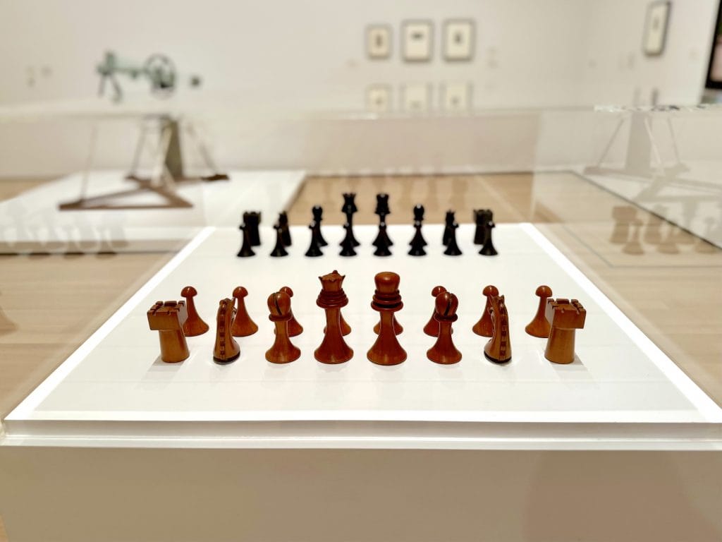 Hand-carved wooden chess pieces designed by Marcel Duchamp, arranged on a white museum plinth with other artworks visible in the blurred gallery background.