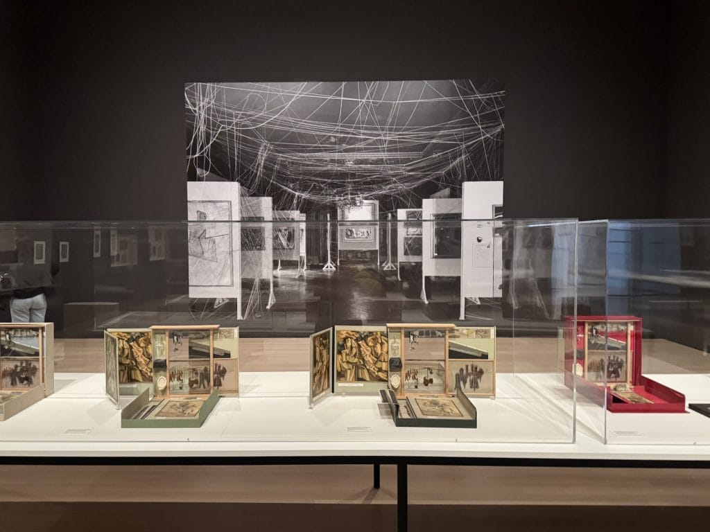 Museum display of Marcel Duchamp’s Boîte-en-valise sets in glass cases, positioned before a large black-and-white photograph of the 1942 "First Papers of Surrealism" exhibition.