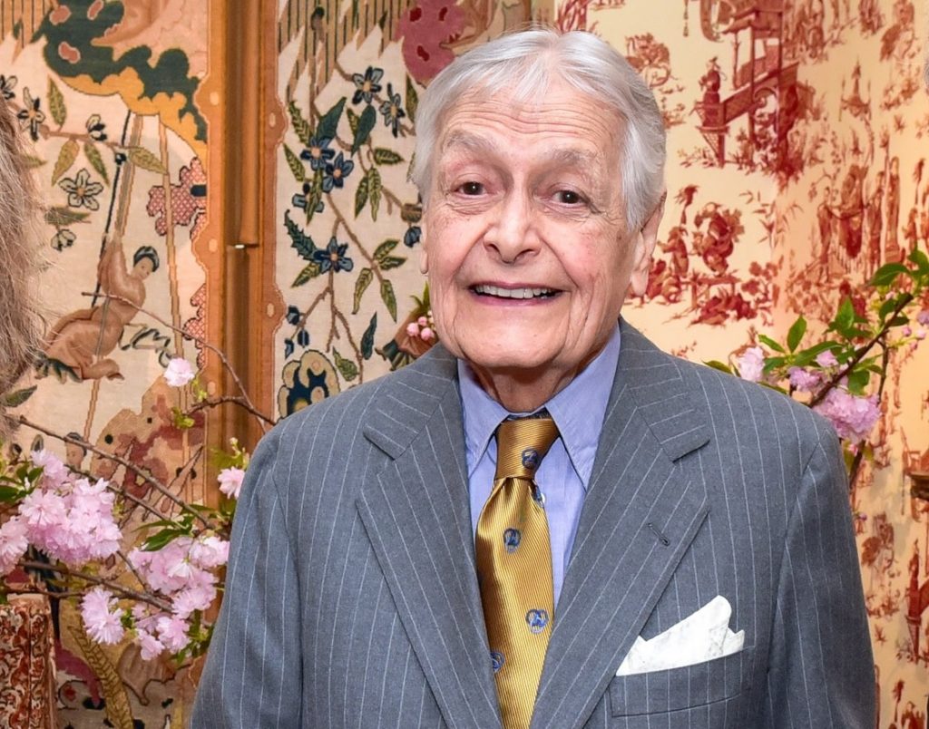 A photo of an older white man with short white hair, smiling warmly while dressed in a gray pinstriped suit, blue shirt, and gold patterned tie. He stands in front of richly patterned wall coverings and floral décor, creating an elegant, traditional backdrop.