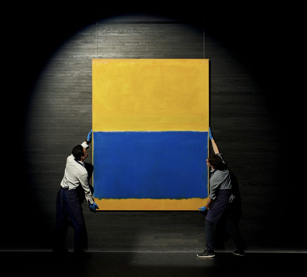 Two art handlers carry a large abstract canvas of bright yellow and deep blue
