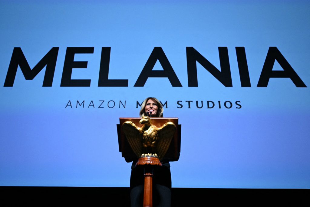 Melania Trump stands behind a golden eagle lectern. A large screen behind her displays "MELANIA" and "AMAZON MGM STUDIOS" in bold black text against blue.