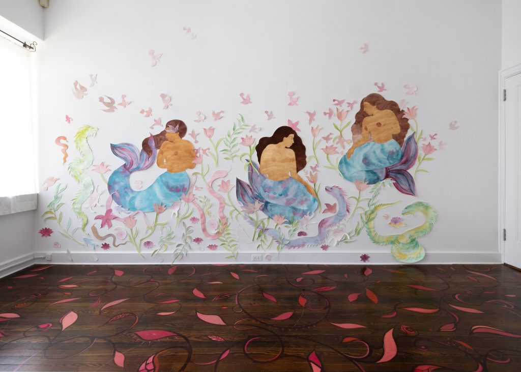 large cut-outs of mermaids on a white wall. the mermaids have long brown hair and light brown skin with blue and purple tails. they are surrounded by pink flowers and dragon-like serpents. 