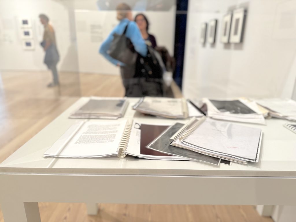 An installation view of Mo Costello’s Untitled (Readers) at the Whitney Biennial, showing several spiral-bound notebooks and papers laid out on a white pedestal.