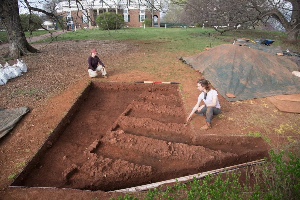 A photograph of two archaeologists crouching around the rectangular, indented remains of an old kiln in a dirt patch before the Monticello main home