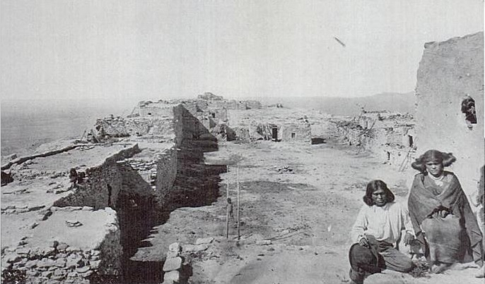 Black-and-white photograph of a desert settlement with partially ruined stone or adobe structures stretching into the distance. In the foreground, two Indigenous individuals sit near a wall on the right side—one wrapped in a blanket, the other wearing a light shirt and pants. The landscape is dry and sparse, with low buildings and open ground extending toward the horizon.