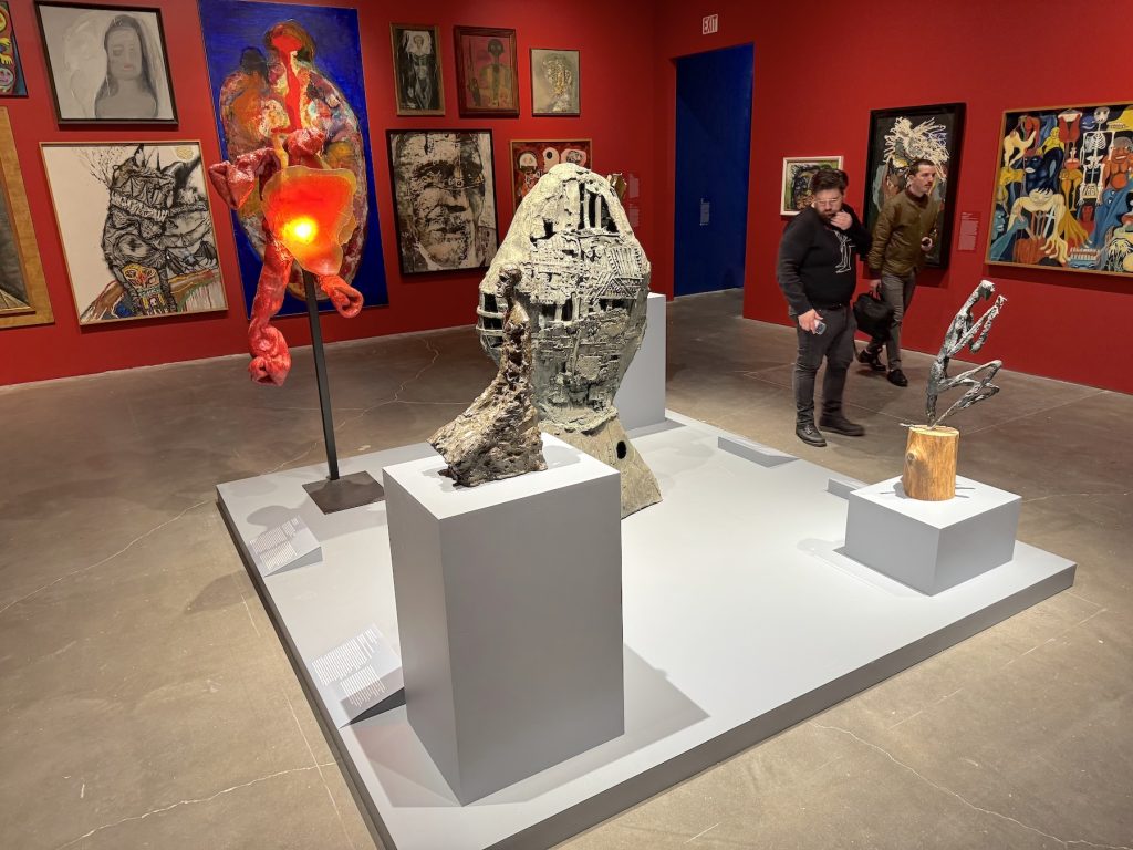 A crowded gallery at the New Museum features various expressive sculptures on a low gray platform against vibrant red walls filled with numerous figurative paintings.