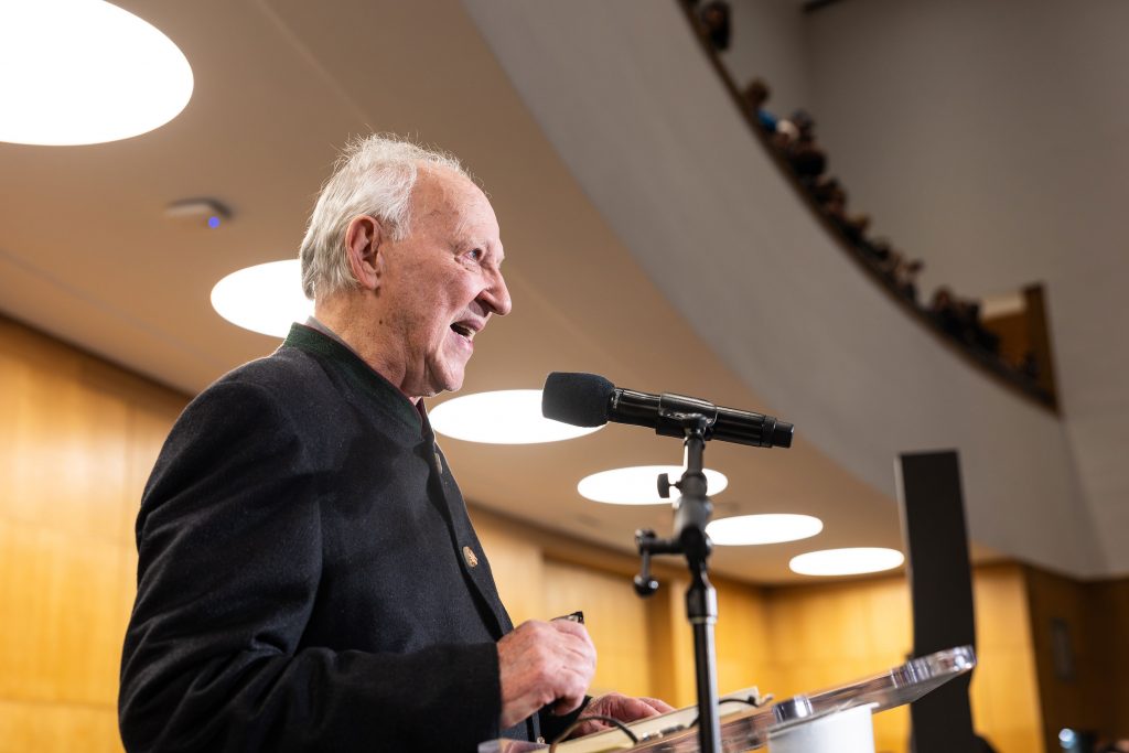 Werner Herzog speaks at podium into microphone inside modern auditorium with curved balcony seating.
