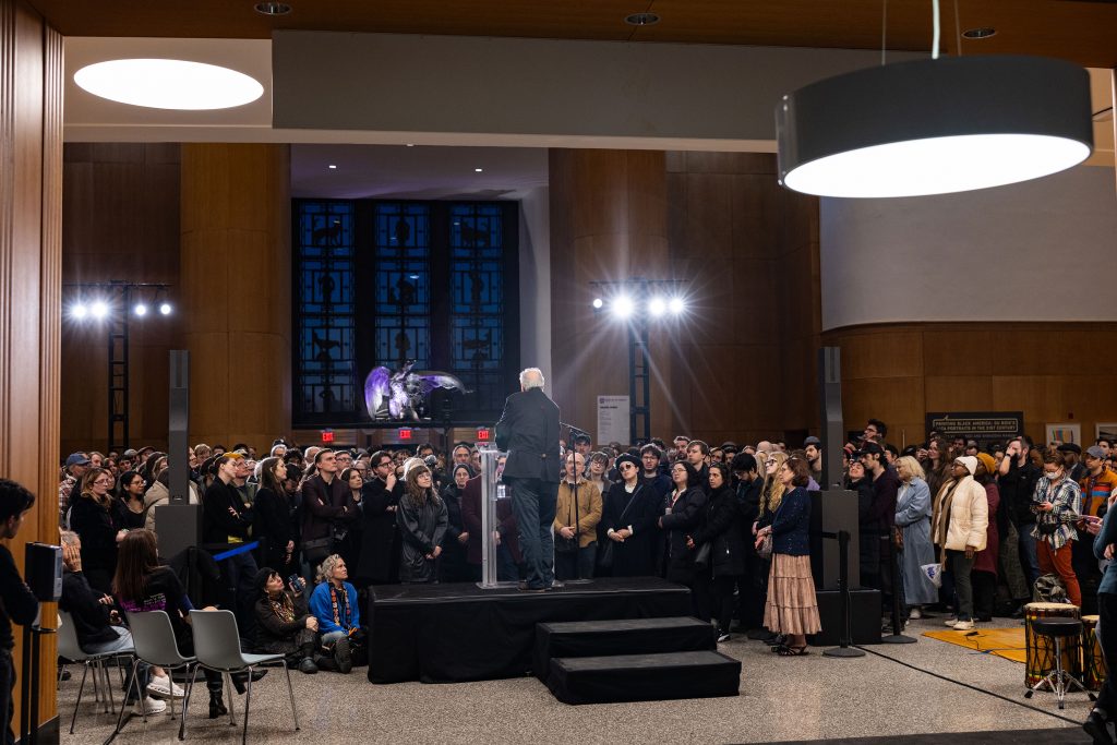 Werner Herzog addresses attentive audience from small stage surrounded by standing crowd in library interior.