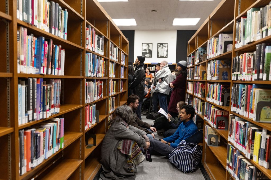 People sit between tall library bookshelves reading and waiting in crowded aisle space together.