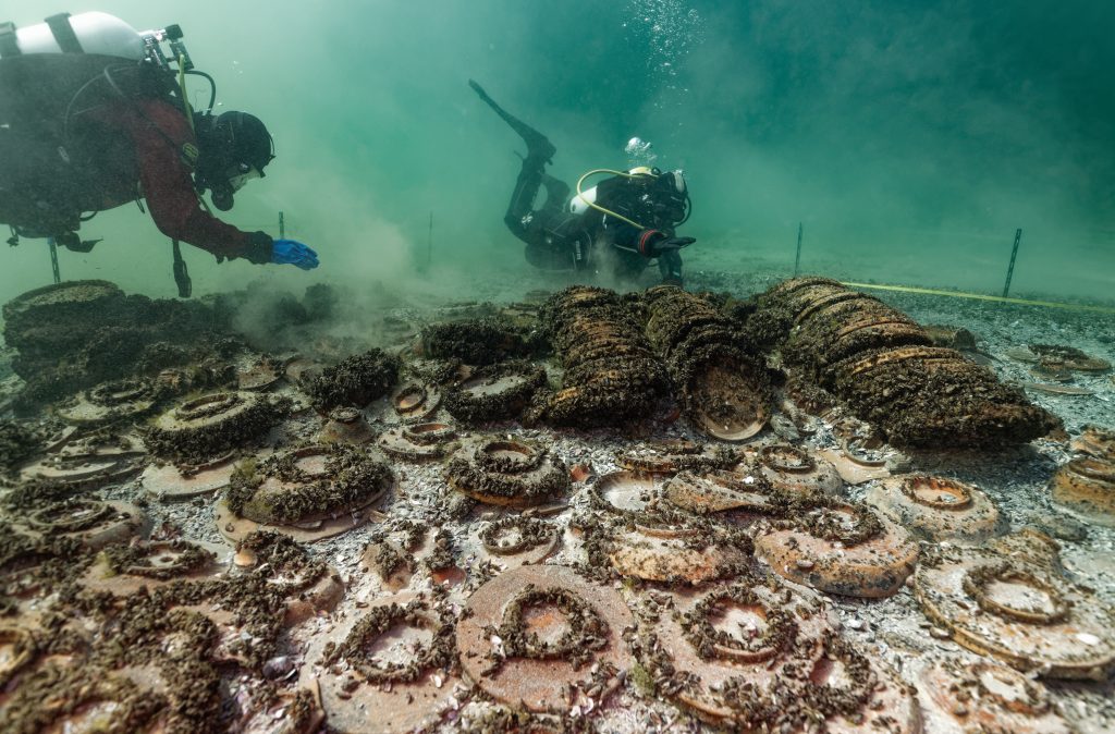 A photograph of two divers exploring a large cluster of old ceramic plates covered with barnacles underwater