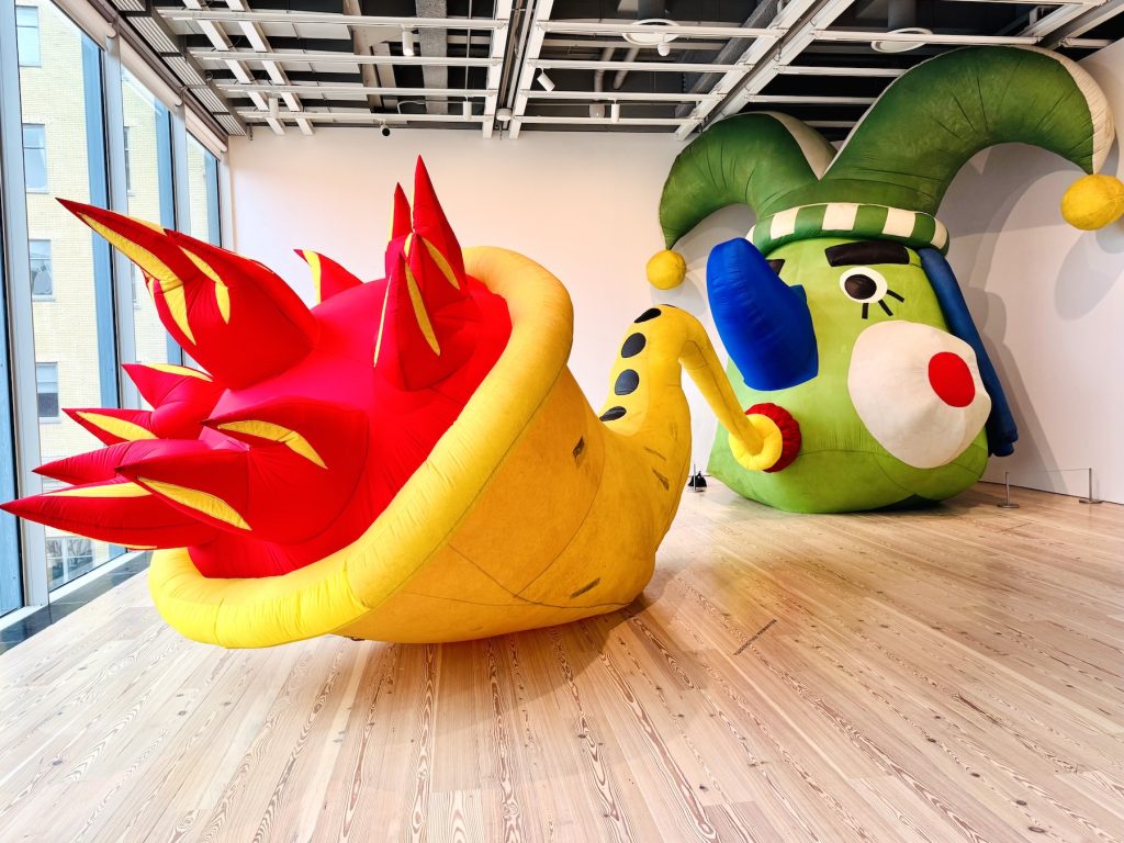 Large-scale, colorful inflatable sculptures by Pat Oleszko in a gallery, featuring a giant green jester head connected by a yellow tube to a spiked horn