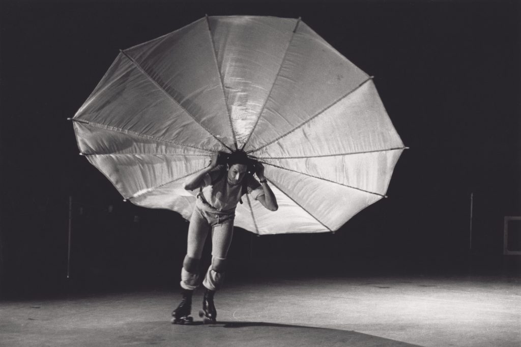 A photograph of Robert Rauschenberg crouched over a bit rollerskating in a vast open rink with a parachute splayed out behind him