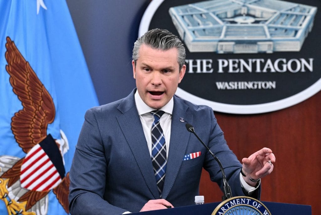 Pete Hegseth speaks at a podium in front of a blue flag and the Pentagon seal. He wears a blue suit and a patterned tie.