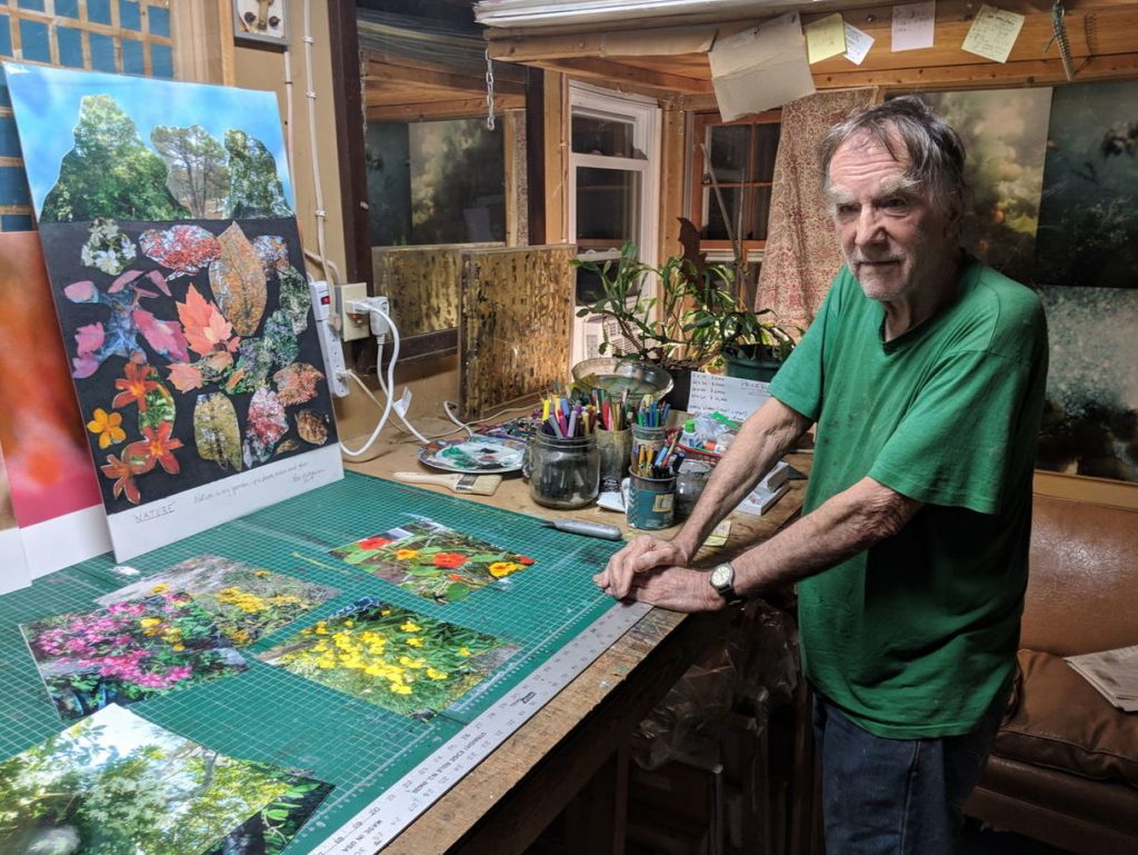 Older man in a green shirt standing at a worktable covered with colorful botanical photographs and art supplies inside a cluttered studio