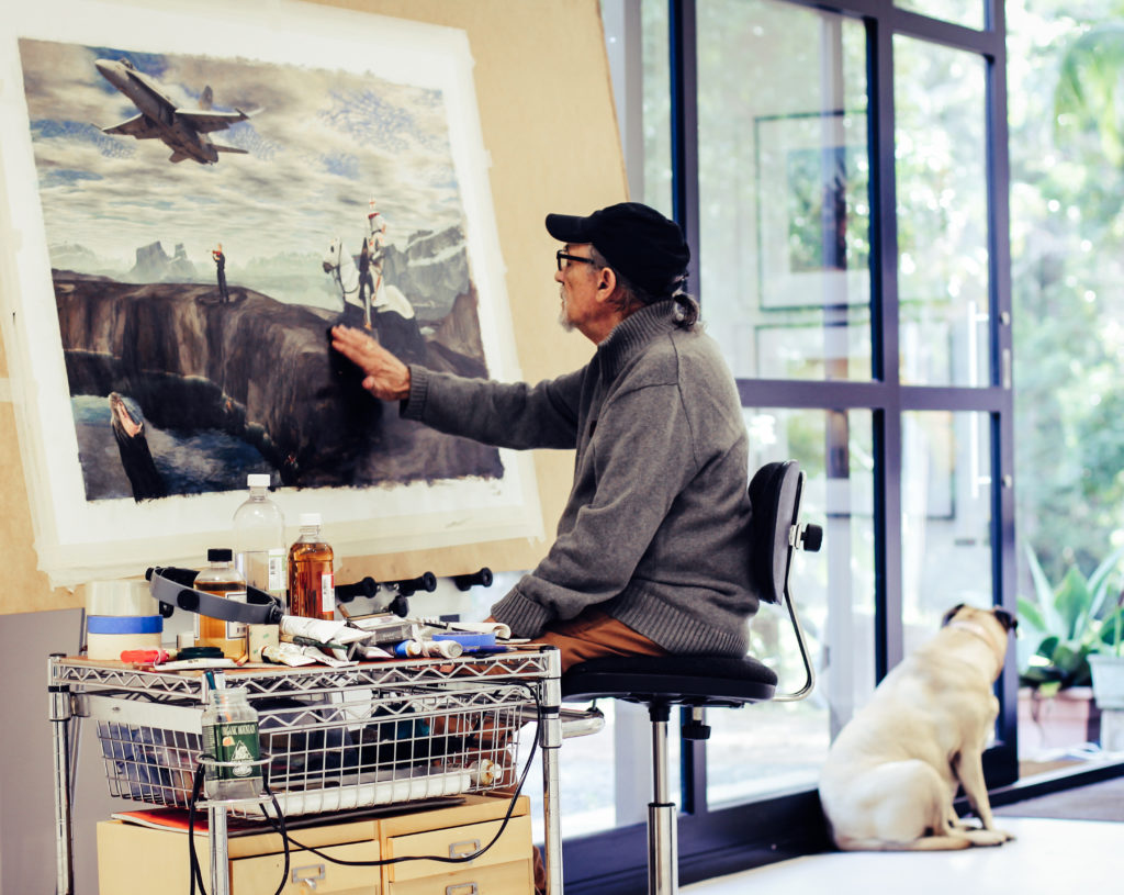 A photo of an older man with a gray beard, glasses, and a black cap sitting on a tall stool in a bright studio, working on a large painting taped to the wall. He wears a gray turtleneck sweater and reaches out to touch the artwork, which depicts a dramatic landscape with figures and a fighter jet. Art supplies are spread across a rolling cart beside him, and a light-colored dog sits on the floor near a glass door, looking outside.