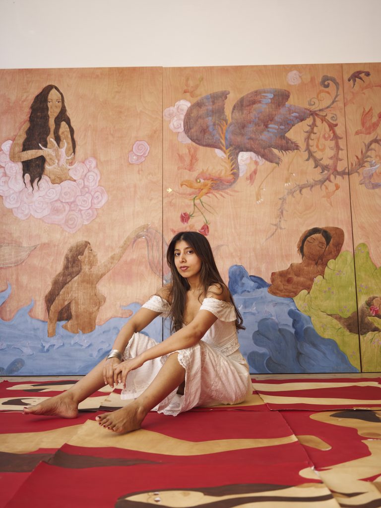 A woman in a white off-the-shoulder dress sits barefoot on a painted floor in front of a large multi-panel mural depicting mermaids, birds, and flowing water in soft pink, blue, and green tones.