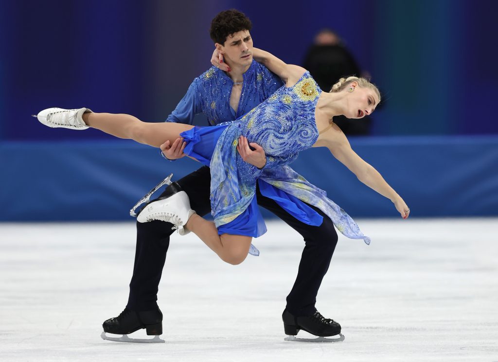 A photo of a white male and a white female figure skating pair performing on an ice rink, with the dark-haired man in a blue embellished costume supporting the blonde woman in a sparkling blue dress as she leans back in a dramatic lift, her leg extended straight out and both wearing white figure skates against a deep blue arena backdrop.
