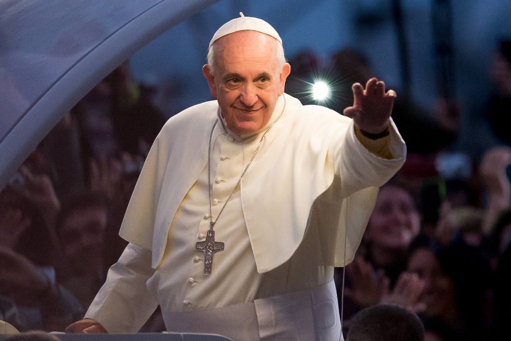 Pope Francis waves to a crowd from a vehicle at dusk, smiling as a flash of light shines behind him. He wears his white papal cassock, cape, and skullcap, with a silver pectoral cross.