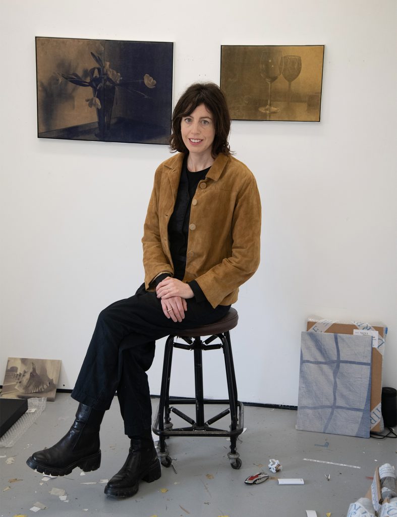 a woman sits cross legged on a stool in a white-walled art studio and looks at the camera, hanging on the wall behind are two silvery toned still lifes that look like old photographs