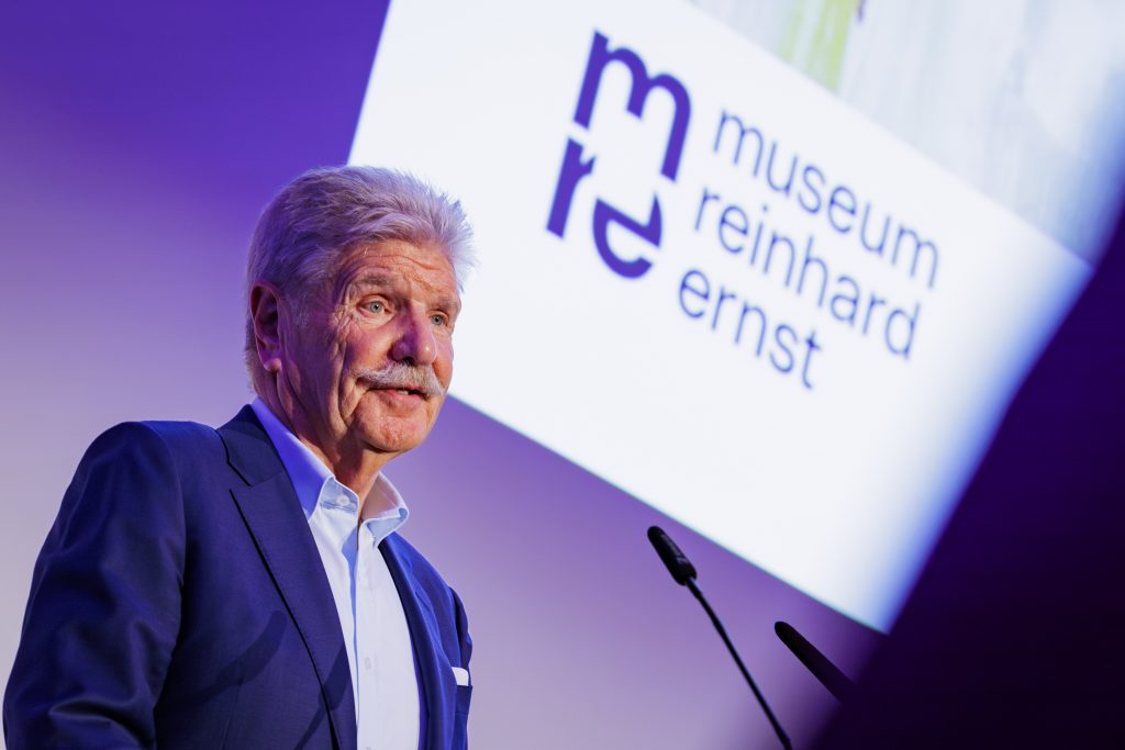 A caucasian man in grey hair and moustache speaking on a podium, against a white backdrop that spells mre Museum Reinhard Ernst