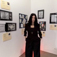 a woman dressed in black stands against a white walled interior on which pieces of art are hung
