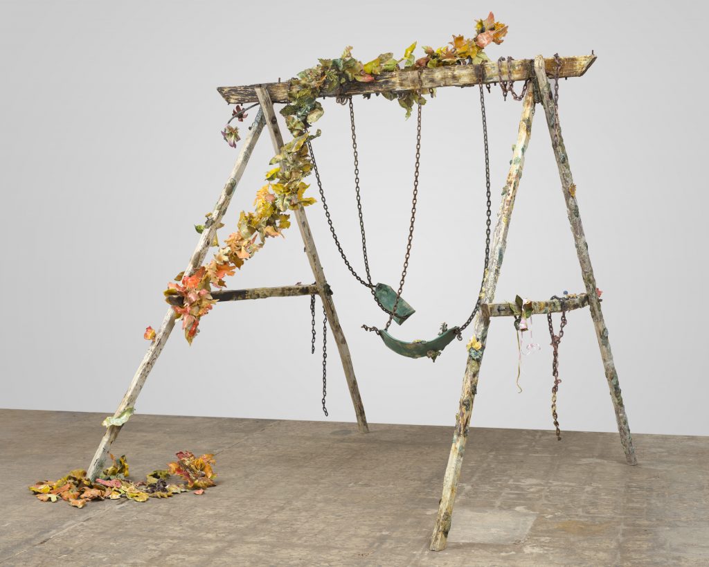 A sculpture by Rebecca Manson in the form of a weathered wooden swing set sculpture with rusted chains and a broken green swing seat, decorated with trailing artificial leaves and flowers in autumn colors, standing on a concrete floor against a plain gray background.