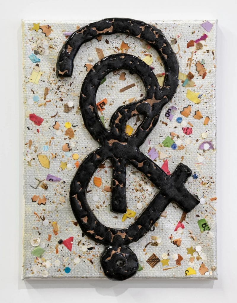 A mixed-media artwork of a thick, black, abstract symbol resembling a looping figure with a cross-like extension, mounted on a white canvas scattered with colorful confetti-like debris and small fragments.